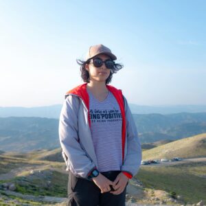 A person in casual attire stands on a mountain with stunning scenic views. Bright daylight conveys a sense of adventure.